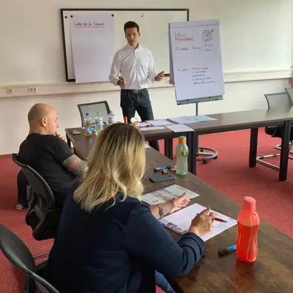 Jörg Vogelgesang leitet ein Verkaufstraining am Flipchart, Teilnehmer arbeiten am Seminartisch mit